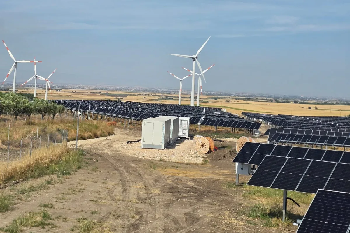 Campo fotovoltaico a terra di grande estensione, con aerogeneratori sullo sfondo